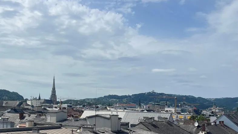 Panoramablick über die Stadt mit historischen Gebäuden und grünen Hügeln im Hintergrund unter bewölktem Himmel.