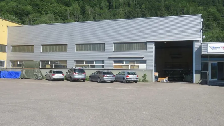 Außenansicht eines Industriegebäudes mit großem Parkplatz und bewaldetem Hügel im Hintergrund.
