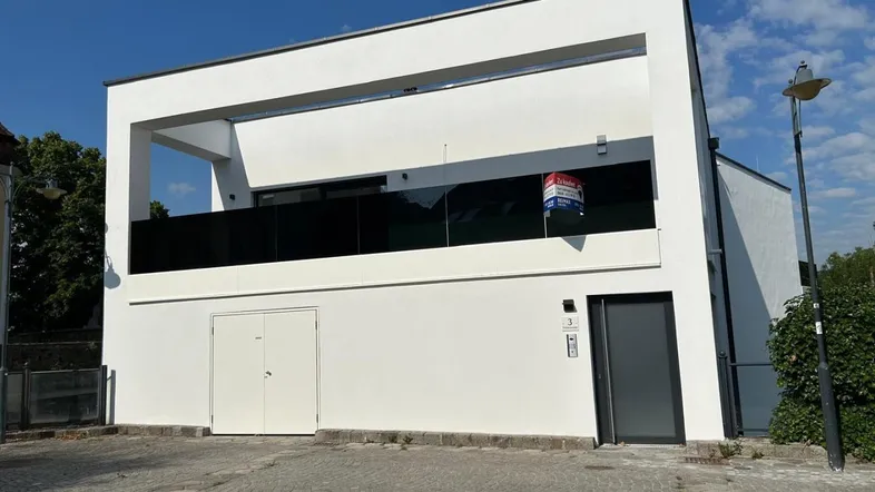 Modernes Gebäude mit weißer Fassade und Balkon, ein Verkaufsschild ist sichtbar.