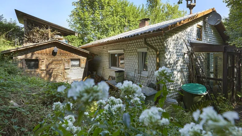Rustikales Haus mit einzigartiger Fassade und angrenzendem Holzschuppen, umgeben von üppigem Grün.