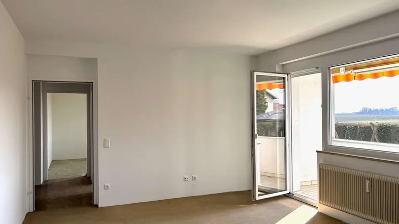 Helles, leeres Zimmer mit Teppichboden, weißen Wänden und Zugang zu einem sonnigen Balkon.