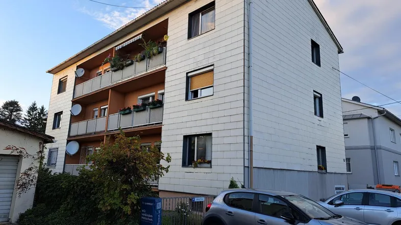 Mehrfamilienhaus mit mehreren Balkonen, weißer Fassadenverkleidung und Satteldach unter blauem Himmel.