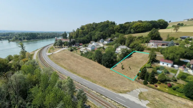Luftaufnahme eines Grundstücks am Flussufer mit angrenzender Straße und Bahnlinie unter klarem Himmel.