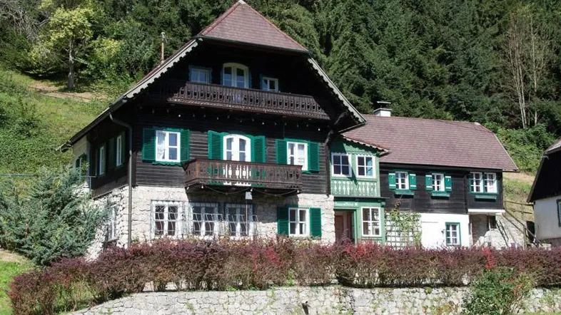Traditionelles alpines Haus mit Steinsockel, Holzfassade, grünen Fensterläden und Balkonen, umgeben von Bäumen.