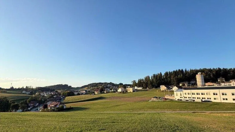Weitläufige grüne Felder mit Blick auf eine ländliche Siedlung und bewaldete Hügel unter klarem Himmel.