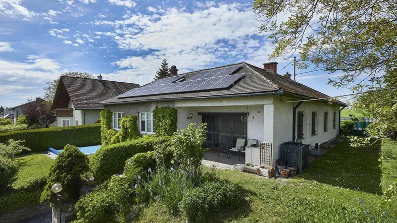 Haus mit Garten, Terrasse und Photovoltaikanlage unter blauem Himmel mit üppiger Vegetation.