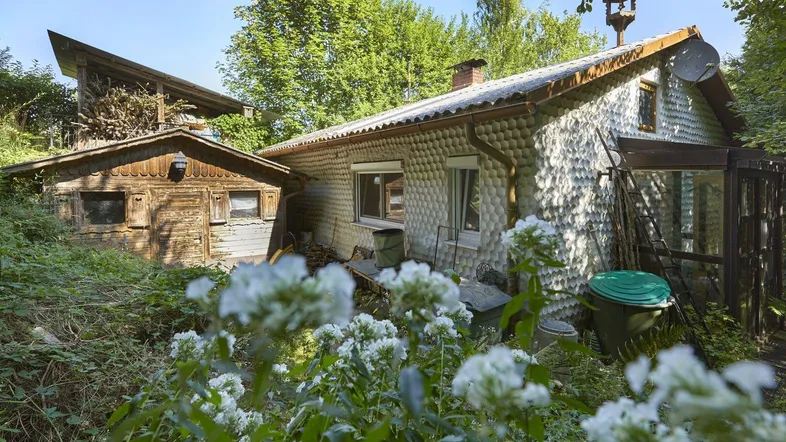 Rustikales Haus mit ungewöhnlicher Fassade und einem Holzschuppen, umgeben von üppigem Grün.