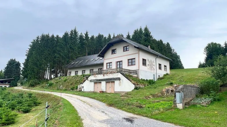 Landhaus auf einem Hügel mit Zufahrtsweg und umgebenden Wäldern unter bewölktem Himmel.