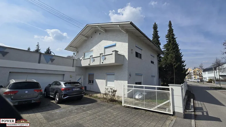 Weißes Einfamilienhaus mit gepflasterter Auffahrt, Garage und zwei geparkten Autos unter blauem Himmel.