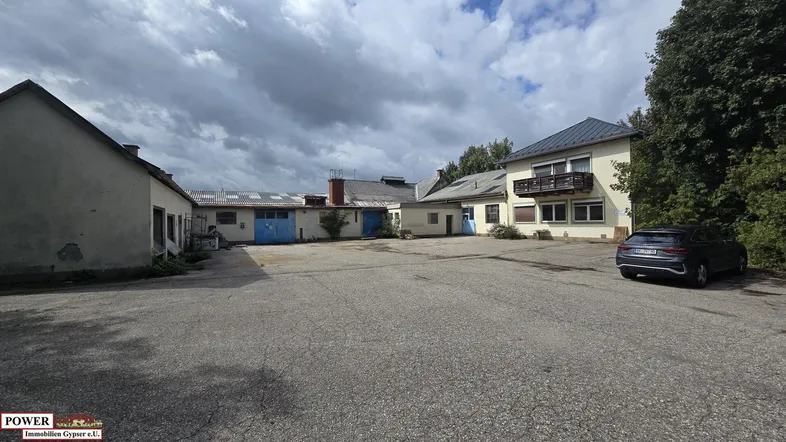 Großer Außenbereich mit mehreren Gebäuden und einem gepflasterten Parkplatz unter bewölktem Himmel.