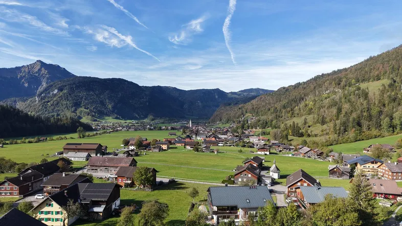 Malerische Luftaufnahme eines Dorfes in einem grünen Tal, umgeben von Bergen unter blauem Himmel.