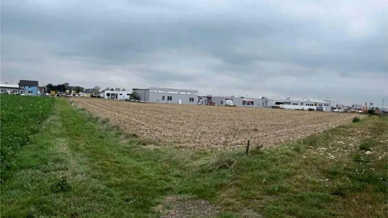 Blick über ein großes Feld mit Gewerbegebäuden im Hintergrund unter einem bewölkten Himmel.