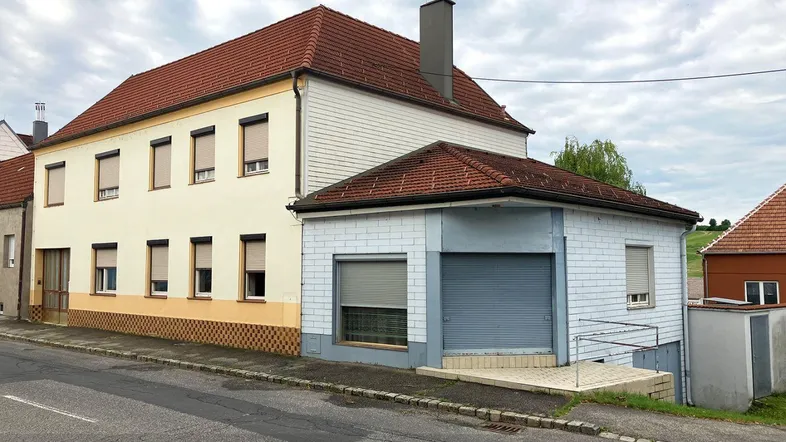 Ansicht des Gebäudes von der Straße mit einem Anbau und Rollläden an den Fenstern.