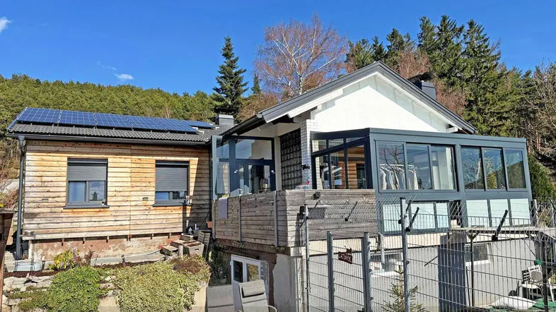 Hausfassade mit integriertem Wintergarten, Solaranlage und kleiner Terrasse im Grünen.