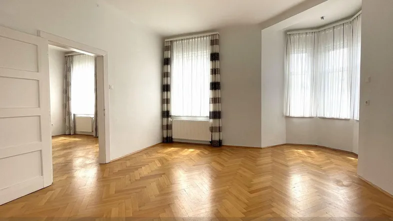 Geräumiges Wohnzimmer mit Fischgrätparkettboden, hohen Decken und einem charmanten Erkerfenster.