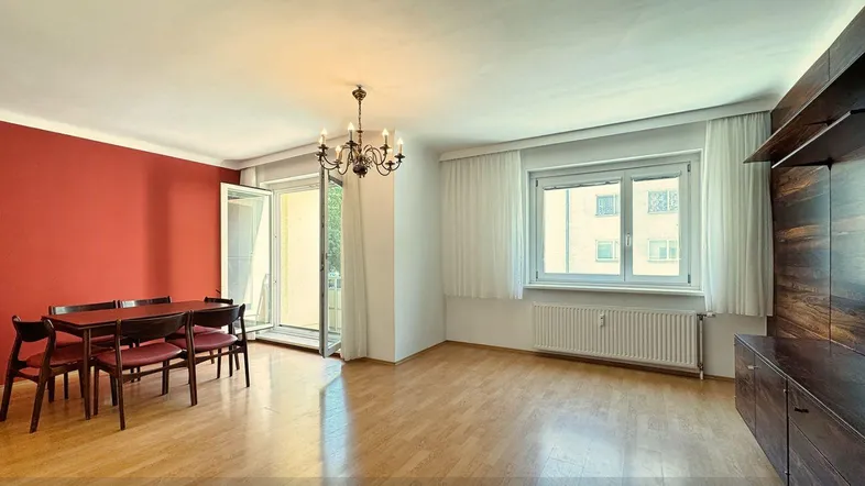 Helles Wohn- und Esszimmer mit Parkettboden, roter Akzentwand und Zugang zum Balkon.