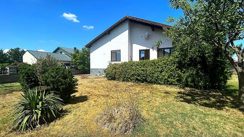 Weißes Einfamilienhaus mit großem Garten und Bäumen unter blauem Himmel.