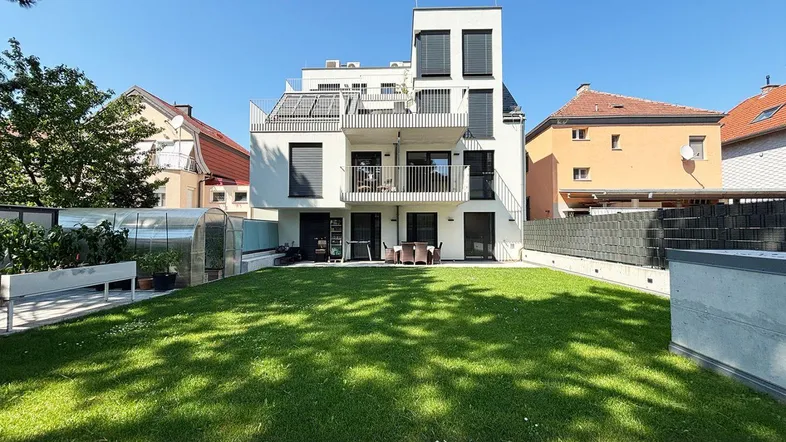 Modernes weißes Gebäude mit mehreren Balkonen, großem Rasen und einem Gewächshaus im Gartenbereich.