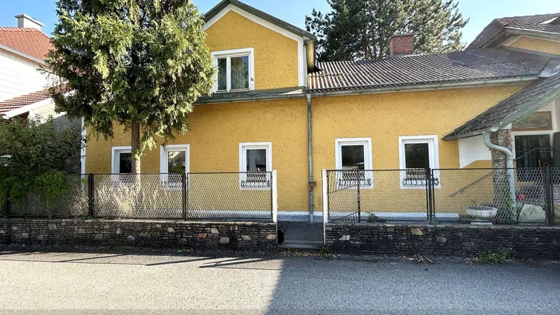 Gelbes Einfamilienhaus mit Zaun und Bäumen im Vorgarten unter blauem Himmel.