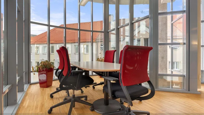 Moderner Konferenzraum mit großen Glasfenstern, Holzboden und roten Bürostühlen, bietet viel Tageslicht.