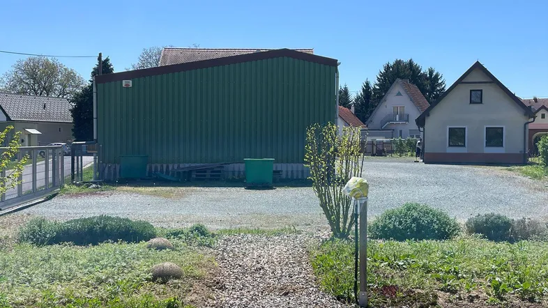 Grünes Lagergebäude auf einem Schotterplatz mit umliegenden Wohnhäusern unter blauem Himmel.