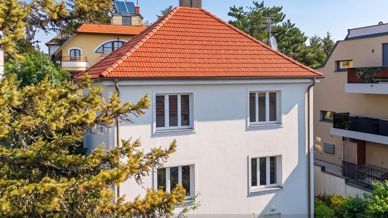 Weißes Haus mit rotem Ziegeldach, umgeben von üppigem Grün und Bäumen, mit modernen Nachbargebäuden.