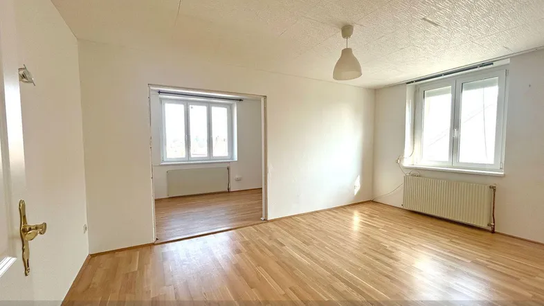 Heller Wohnraum mit Parkettboden und großen Fenstern, offen zu einem weiteren Zimmer.