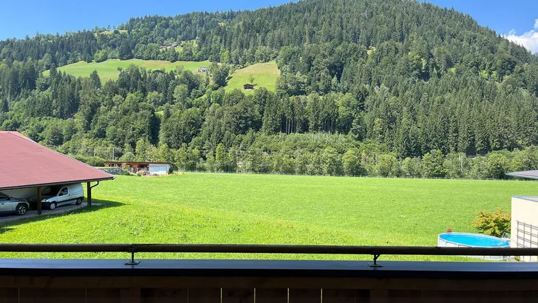 Panoramablick von einem Balkon über grüne Wiesen und bewaldete Berge unter blauem Himmel.