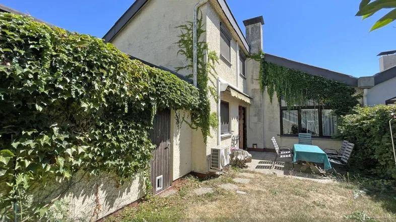 Hausfassade mit bewachsenen Wänden, kleiner Terrasse und Gartenbereich unter blauem Himmel.