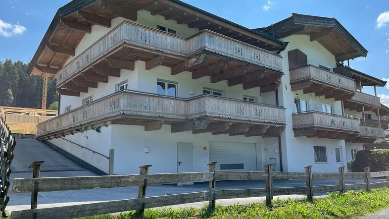 Alpines Gebäude mit mehreren Holzbalkonen, Garagenzugang und umgeben von grüner Landschaft.