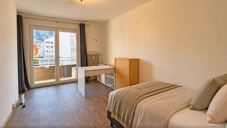 Helles Schlafzimmer mit Holzboden, Schreibtisch und direktem Zugang zu einem Balkon.