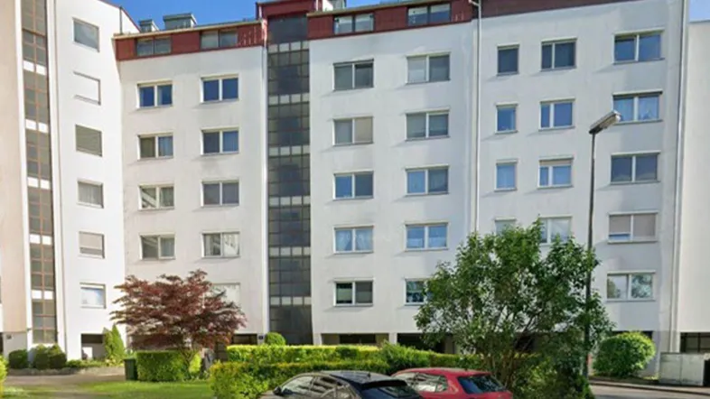 Mehrstöckiges Wohngebäude mit weißer Fassade, umgeben von Grünflächen und parkenden Autos.