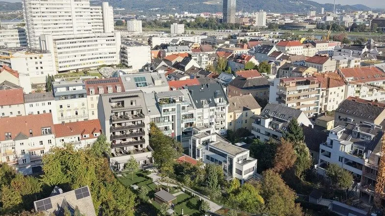 Stadtansicht mit Wohngebäuden, viel Grün und Bergen im Hintergrund unter blauem Himmel.