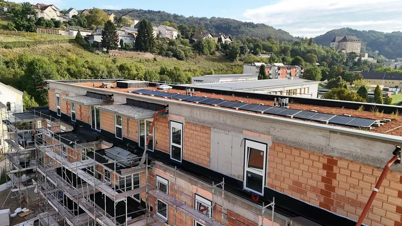Neubau im Bau mit Gerüst, Ziegelwänden und Solarpaneelen auf dem Dach, umgeben von grünen Hügeln.