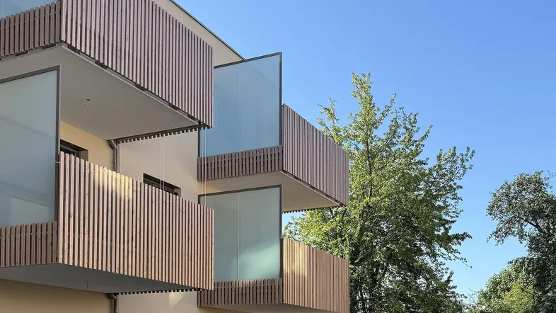 Moderne Gebäudefassade mit mehreren Balkonen, ausgestattet mit Holzlamellen und Glasgeländern, umgeben von Grün.