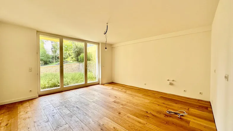Helles Zimmer mit Holzboden und großen Fenstern, die viel Tageslicht hereinlassen und einen Blick ins Grüne bieten.