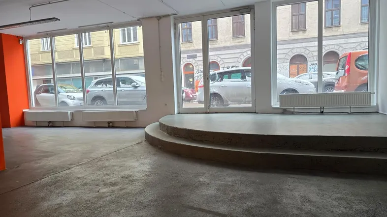 Großer, offener Gewerberaum mit Schaufenstern zur Straße und einer erhöhten Plattform im Vordergrund.