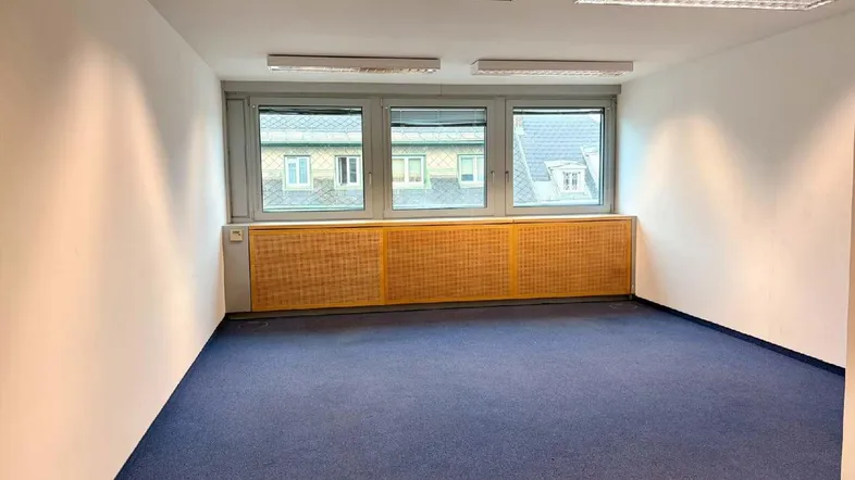 Leerer Büroraum mit blauem Teppichboden, großen Fenstern und Deckenbeleuchtung, bereit zur Nutzung.