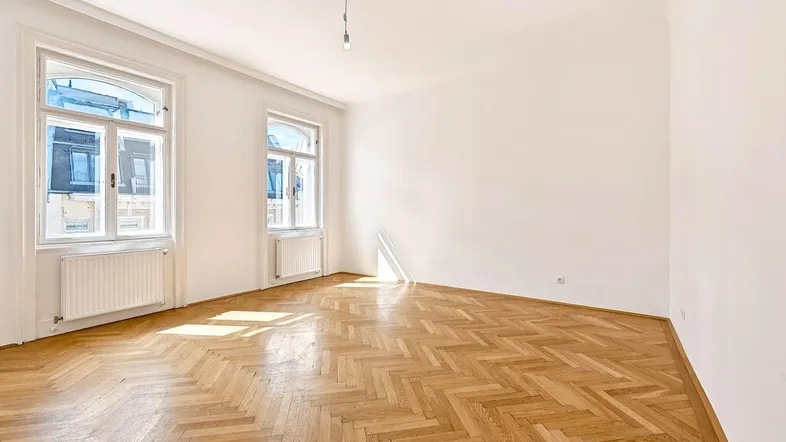 Helles Zimmer mit Fischgrätparkett und zwei großen Fenstern, das viel Tageslicht hereinlässt.