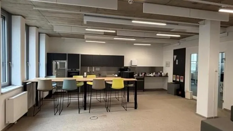 Moderne Büroküche mit langer Theke, bunten Hockern, dunklen Schränken und freiliegender Betondecke.