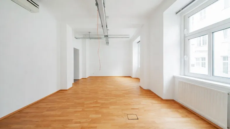 Langer, heller Büroraum mit Holzboden, Klimaanlage und großen Fenstern, die viel Tageslicht hereinlassen.