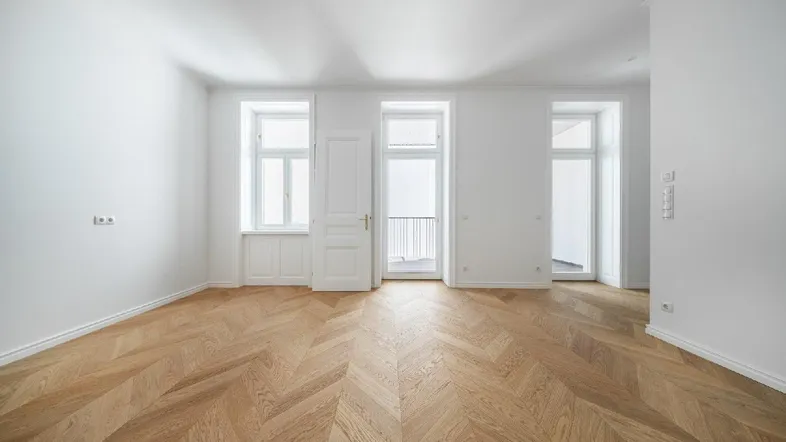 Helles, leeres Zimmer mit Fischgrätparkettboden und Zugang zu einem französischen Balkon.