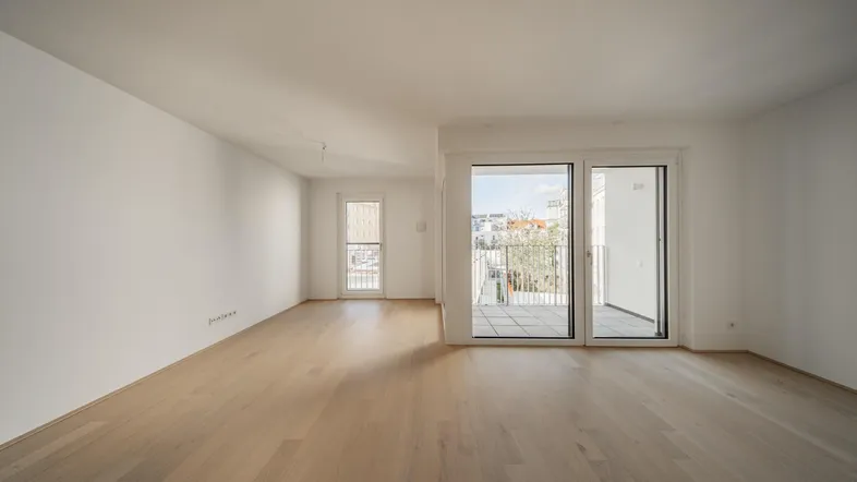 Helles, leeres Zimmer mit Parkettboden und Zugang zu einem Balkon oder einer Terrasse.