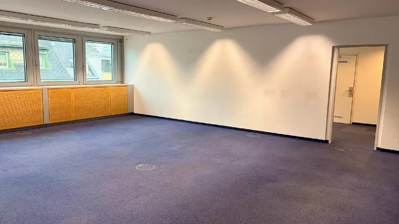 Geräumiger Büroraum mit blauem Teppichboden, hellen Wänden und Zugang zu weiteren Bereichen im Gebäude.
