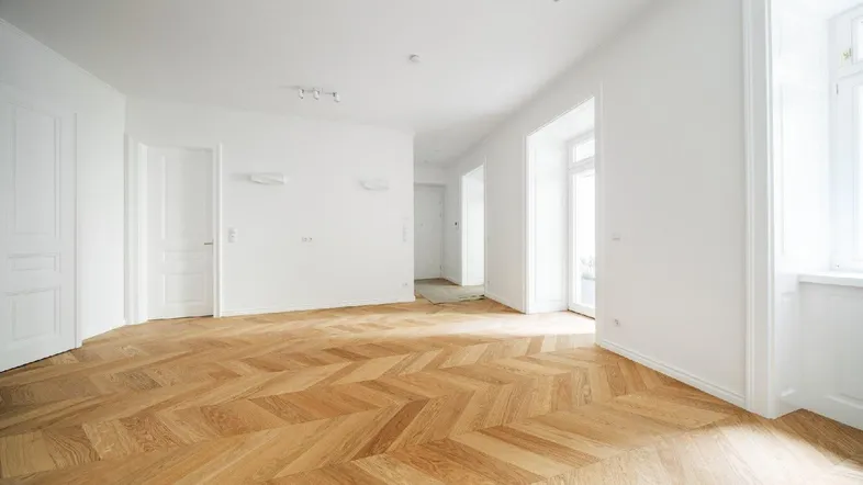 Helles Zimmer mit elegantem Fischgrätparkett, weißen Wänden und Zugang zu weiteren Räumen.