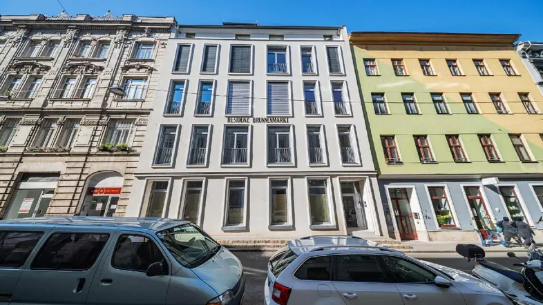 Moderne Fassade eines Neubaus an einer belebten Stadtstraße mit parkenden Autos.
