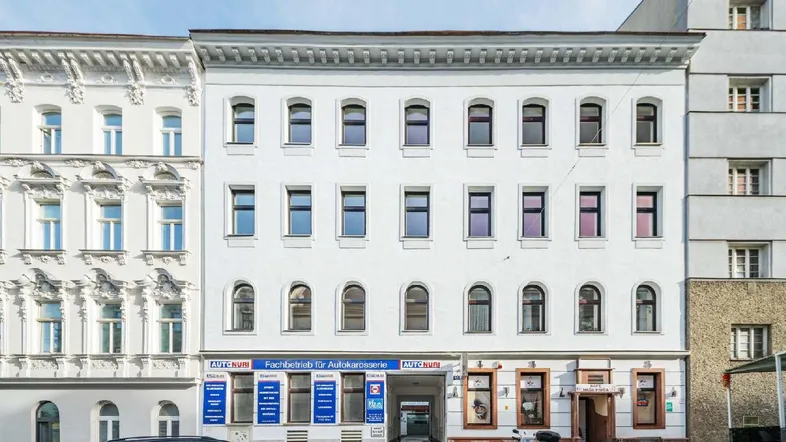Die weiße Fassade des Mehrfamilienhauses mit vielen Fenstern und Geschäften im Erdgeschoss.
