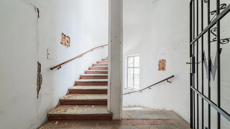 Historisches Treppenhaus mit abgenutzten Stufen, Fenster und Gittertor, das natürliches Licht hereinlässt.