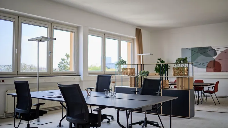 Heller, moderner Büroraum mit großen Fenstern, mehreren Arbeitsplätzen und minimalistischer Einrichtung.