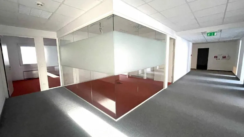 Geräumiges Büro mit Teppichboden und modernen Glaswänden, die separate Arbeitsbereiche schaffen. Helle Beleuchtung durch Deckenleuchten.